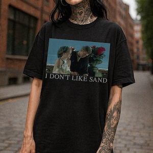 May include: Black t-shirt featuring a movie scene with the text "I DON'T LIKE SAND". The graphic shows two people sitting near a flower pot. The shirt is made of a soft material.