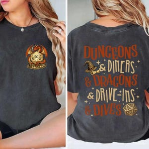 May include: Dark grey t-shirt featuring a dragon and dice graphic on the front. The back of the shirt displays text that reads "Dungeons & Diners & Dragons & Drive-Ins & Dives" in orange and white. The shirt is made of a soft material.