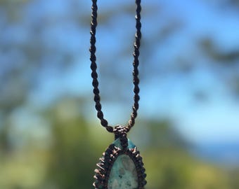 May include: A handcrafted macrame necklace featuring a teardrop-shaped pendant. The pendant showcases a light blue and green stone, encased in a brown macrame weave. The necklace cord is a twisted brown rope, perfect for a bohemian style.