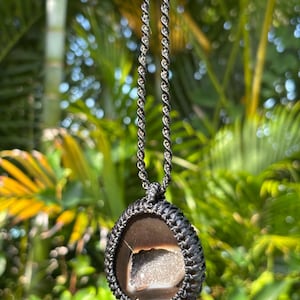 Puede incluir: Un collar artesanal con un colgante de piedra marrón oscuro, más claro en el interior, engastado en un tejido de macramé negro. El colgante cuelga de un cordón de macramé negro a juego, ofreciendo un aspecto natural y terroso.