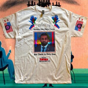 Könnte beinhalten: Cremefarbenes T-Shirt mit dem Text "Anxiety Has Many Faces But There Is Only One". Das Shirt zeigt ein Porträt, farbenfrohe Vogelgrafiken und eine Werbung für ein verschreibungspflichtiges Medikament. Die Ärmel haben Flaggen-Grafiken.