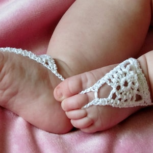 May include: A pair of white crocheted baby booties with a delicate floral design. The booties are worn on a baby's feet, resting on a pink blanket.