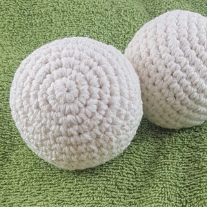 May include: Two off-white, crocheted balls sit on a green, textured surface. The balls are round, with visible stitch patterns creating a spiral design. The texture of the balls contrasts with the background.