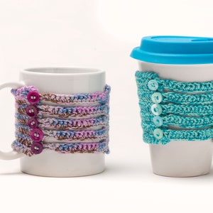 May include: Two white ceramic mugs with crocheted covers. The mug on the left has a purple, blue, and brown crocheted cover with pink buttons. The mug on the right has a blue crocheted cover with white buttons.