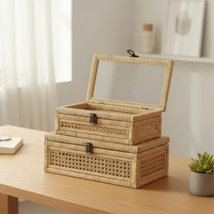 May include: Two stacked, rectangular woven boxes with hinged lids. The top box has a clear glass lid, and both have metal clasps. The boxes are made of light brown woven material and sit on a wooden surface. The bottom box has a decorative woven pattern.