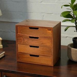 May include: A small, four-drawer wooden storage chest. The chest is made of light brown wood with a natural finish. Each drawer has a simple, recessed handle. The chest is sitting on a dark brown surface.