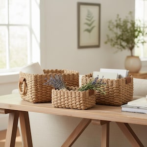 May include: Three woven storage baskets in varying sizes. The baskets are a natural tan color and have a scalloped edge. One basket has a leather handle, and the others have leather accents. The baskets are on a wooden table and contain items.