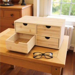 May include: A light-colored bamboo desk organizer with four drawers, one of which is open. The organizer sits on a wooden table, with a pair of black-framed eyeglasses resting on the table. The drawers have curved cutouts for handles.