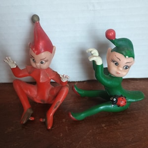 May include: Two vintage elf figurines. One elf is in a red outfit and hat, the other in green. Both have painted faces and are in a seated position. A ladybug is on the green elf's leg.