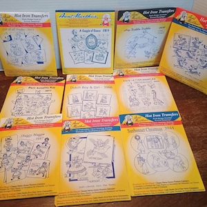 May include: A collection of vintage Aunt Martha's Hot Iron Transfers, featuring various designs for embroidery, fabric painting, and quilting. The packets are yellow with illustrations and text, including titles like "Cat of all Trades" and "Sunbonnet Christmas."