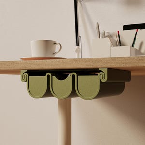 Puede incluir: Un organizador verde debajo del escritorio con tres compartimentos curvos. El organizador está montado debajo de un escritorio de madera, con una taza de café blanca y un platillo visibles en la superficie del escritorio. También hay una lámpara de escritorio y útiles de oficina.