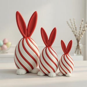 May include: Three decorative Easter bunny figurines. Each bunny has a white body with red stripes, red ears, and white feet. The bunnies are arranged in a row, decreasing in size from left to right. A bowl of pastel-colored eggs is in the background.