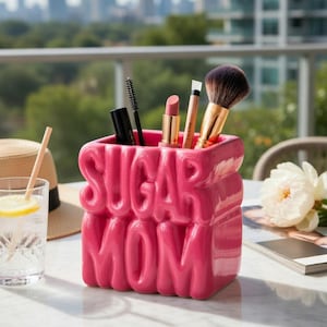 May include: A vibrant pink ceramic pot, embossed with the words "SUGAR MOM", filled with makeup brushes and lipsticks. The pot is on a marble table, with a city skyline in the background. A glass of water and a straw are also visible.