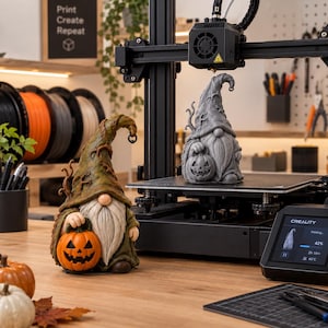 May include: A 3D printer is actively printing a grey gnome figurine holding a pumpkin. Beside it sits a finished, hand-painted gnome with a green hat and orange pumpkin. The printer screen displays a 42% progress bar and the brand name Creality.