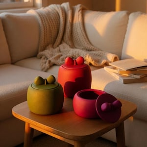 Puede incluir: Tres recipientes de cerámica coloridos con tapas, cada uno con forma redondeada y un asa en forma de lazo. Los recipientes son de color verde, rojo y magenta, colocados sobre una mesa de madera. Un sofá blanco y una manta beige en el fondo.