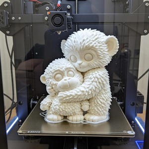 May include: A 3D-printed sculpture of two white monkey-like figures embracing. The larger figure hugs the smaller one. The figures are on a 3D printer platform. The printer has a black frame and the text "3D PRINT MATERIAL".