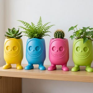 Pode incluir: Quatro vasos coloridos e extravagantes em amarelo, azul, rosa e verde, cada um com um design de rosto sorridente e pés pequenos. Os vasos contêm várias plantas, incluindo uma suculenta, uma samambaia, um cacto e uma trepadeira, expostos numa prateleira de madeira.