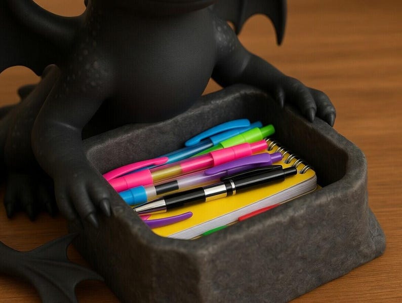 Puede incluir: Un organizador de escritorio negro con forma de drag&oacute;n contiene bol&iacute;grafos de colores y un bloc de notas amarillo. El organizador est&aacute; hecho de un material gris oscuro y se encuentra sobre una superficie de madera. Los bol&iacute;grafos son de varios colores, incluyendo rosa, azul, verde y morado.