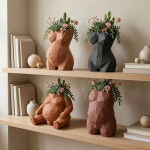 May include: Four geometric torso-shaped planters in terracotta, black, and reddish-brown hues. Two planters are standing and two are sitting, each featuring a faceted surface texture. Each planter contains a variety of green succulents and small pink flowers.