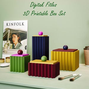 Könnte beinhalten: Eine Sammlung farbenfroher, geometrischer 3D-Druck-Box-Sets. Die Boxen sind in Grün-, Burgunder-, Gelb- und Blautönen gehalten und haben jeweils eine kontrastierende Kugel. Das Bild zeigt auch ein Kinfolk-Magazin und Pinsel.