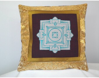 May include: A square cushion with a gold embroidered border and a blue and white mandala design in the center. The cushion is made of a dark brown fabric.