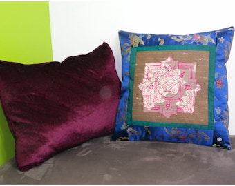May include: Two decorative throw pillows. One pillow is a deep burgundy velvet with a smooth texture. The other pillow is blue with a floral pattern and a brown center panel with a pink and white mandala design.