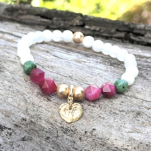 Puede incluir: Una pulsera de cuentas con una serie de cuentas blancas redondas y lisas, acentuadas con dos cuentas verdes moteadas, cuatro cuentas magenta facetadas y dos cuentas doradas metálicas. Un dije dorado texturizado en forma de corazón cuelga del centro.