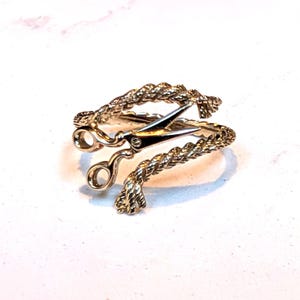 Peut inclure: Une bague en métal doré dotée d'un anneau à la texture de corde torsadée qui s'enroule autour du doigt. L'élément central de la bague est une petite paire de ciseaux ouverts et détaillés, intégrée au design reliant les deux extrémités.