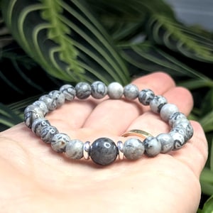 Puede incluir: Pulsera de cuentas grises y blancas con una gran cuenta central gris oscuro. La pulsera presenta cuentas estampadas y lisas, ensartadas en una banda elástica. Acentos metálicos plateados a ambos lados de la cuenta central. Se muestra sobre un fondo verde con hojas. Diámetro de las cuentas: aprox. 0,8 cm.