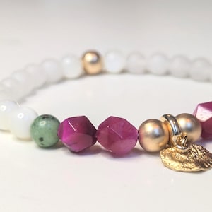 Puede incluir: Una pulsera elástica con cuentas blancas lisas, cuentas magenta facetadas, una cuenta verde moteada y dos cuentas esféricas de color dorado mate. Un pequeño dije de metal dorado texturizado cuelga del centro de la pulsera.