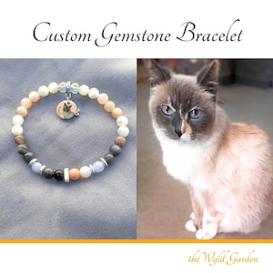 Puede incluir: Una pulsera de cuentas con piedras redondas en tonos blanco, melocotón, gris y azul claro, con espaciadores plateados y un dije circular con un retrato de gato y el texto Pearl. Aparece el texto Custom Gemstone Bracelet y the Wyld Garden.