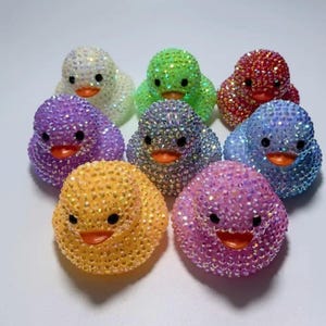 May include: A collection of eight rubber ducks covered in small, reflective, multi-colored rhinestones. Each duck features a bright orange beak and two black eyes, with individual ducks colored in white, green, red, purple, blue, yellow, and pink.
