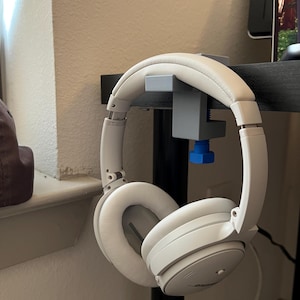 May include: White over-ear headphones hanging from a gray and blue headphone stand. The headphones have a padded headband and earcups. The stand is clamped to a black desk. The headphones are a neutral color.