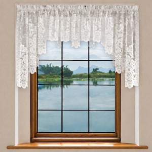 May include: White lace window valance with floral patterns. The valance is draped over a wooden window frame, partially obscuring the view of a lake and mountains. The lace has a scalloped edge.