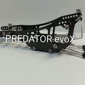 Puede incluir: Dispositivo mecánico negro y plateado con el texto "PREDATOR evoX". El dispositivo tiene múltiples agujeros y varillas de conexión. El dispositivo está sobre una superficie blanca.