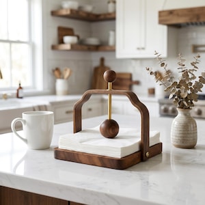 May include: A square wooden napkin holder with a dark brown finish, featuring a central gold-toned metal rod topped with a wooden sphere and a matching weighted wooden ball to secure a stack of white paper napkins in place on a flat base.