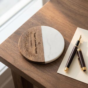 May include: A circular coaster featuring a split design of natural brown wood and white marble. The wood section is engraved with a botanical wreath illustration and the text "the Your Custom Name FAMILY". A dark brown fountain pen lies nearby.