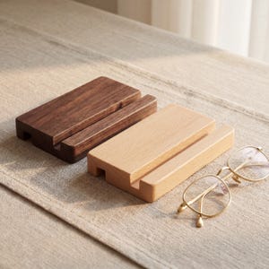 以下が含まれることがあります： 2つの木製電話スタンド。濃い茶色と薄い茶色の2つがニュートラルな表面に置かれています。金縁の眼鏡が近くにあります。スタンドには電話を保持するためのスロットがあります。