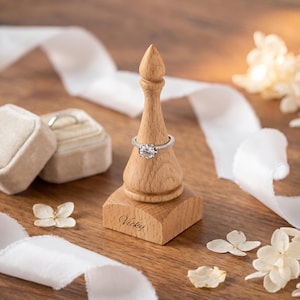 May include: A light-colored wooden ring holder shaped like a chess piece with a pointed top and a square base. The name Vicky is engraved on the front of the base. A silver ring with a round clear stone is placed on the tapered neck of the holder.