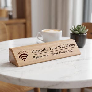 May include: A wooden WiFi sign with the word "welcome" and a WiFi symbol. The sign displays "Network: Your Wifi Name" and "Password: Your Password" in black text. The sign is on a white marble table, with a coffee cup and plant.