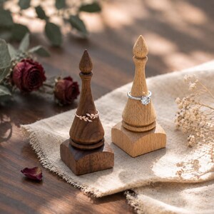 May include: Two wooden ring holders with square bases and tapered, pointed tops. One holder is crafted from dark brown wood and displays a rose gold leaf-patterned ring. The other is made of light tan wood and holds a silver ring with a clear stone.