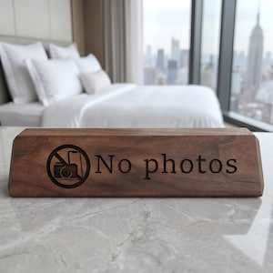 May include: A rectangular wooden sign with the words "No photos" engraved in a serif font. The sign also features a graphic of a camera with a line through it, indicating photography is prohibited.