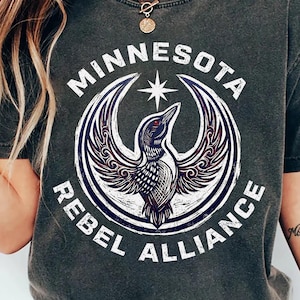 May include: Dark gray t-shirt featuring a circular design with a bird and the words "MINNESOTA REBEL ALLIANCE". The design is in white and purple. The shirt has short sleeves and a crew neck.