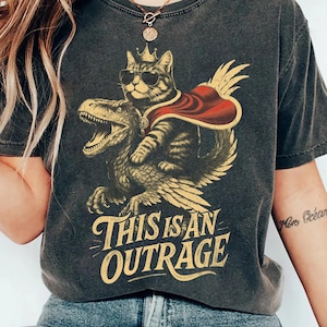 May include: Dark gray t-shirt featuring a cat wearing sunglasses and a crown, riding a dinosaur with a red cape. The text "THIS IS AN OUTRAGE" is printed in a vintage style. The shirt is a relaxed fit.