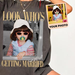 May include: A charcoal gray oversized t-shirt featuring a large graphic print of a child wearing a white bucket hat, sunglasses, and a pacifier. The text reads LOOK WHO'S GETTING MARRIED and Sophia. A small inset photo shows the same child.