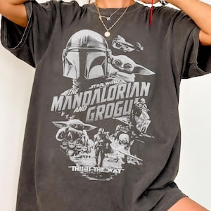 May include: Dark gray t-shirt featuring a graphic from the Star Wars series, "The Mandalorian and Grogu." The design includes images of the Mandalorian, Grogu, and other characters, with the phrase "THIS IS THE WAY" at the bottom.