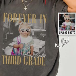 Op de afbeelding: Een antracietgrijs T-shirt met korte mouwen en een grote centrale afbeelding van een kind met een hartvormige zonnebril en een bloemenjurk. De tekst luidt FOREVER IN THIRD GRADE en Mrs. Rachel. Een kleine inzetfoto toont het kind.