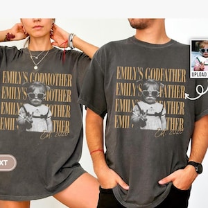 Könnte beinhalten: Ein anthrazitfarbenes Kurzarm-T-Shirt mit einem Grafikdruck eines Kindes, das eine herzförmige Sonnenbrille trägt. Der Text "EMILY'S GODMOTHER" oder "EMILY'S GODFATHER" ist in goldener Schrift wiederholt, mit "Est. 2020" darunter gedruckt.