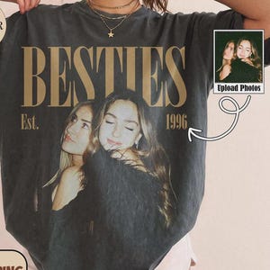 Könnte beinhalten: Ein anthrazitfarbenes Kurzarm-T-Shirt mit einem großen Grafikdruck von zwei sich umarmenden Personen. Der Text BESTIES ist in großen goldenen Buchstaben über dem Bild gedruckt, mit Est. 1996 auf beiden Seiten. Das Shirt hat einen Rundhalsausschnitt.