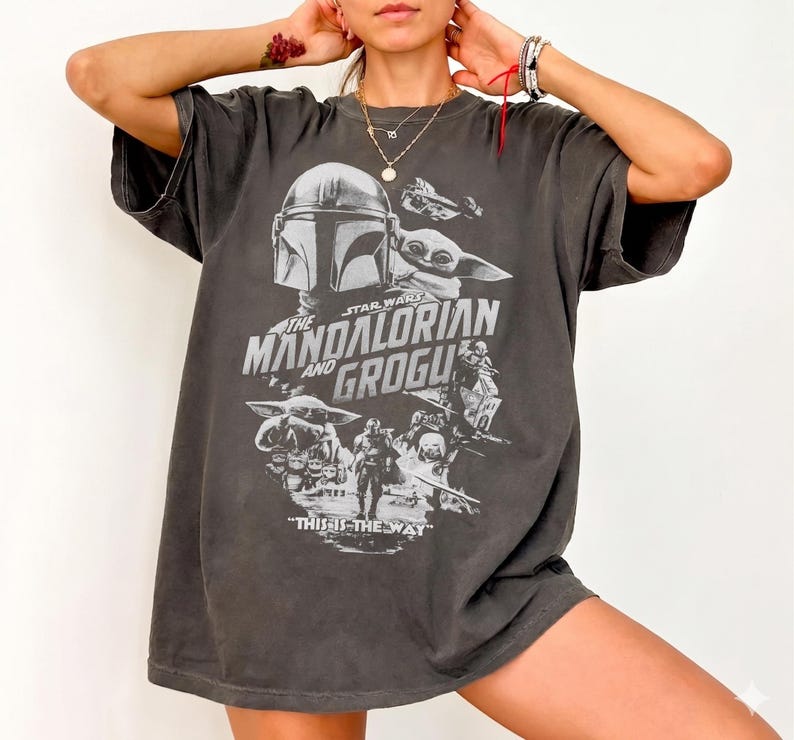 May include: Dark gray t-shirt featuring a graphic from the Star Wars series, "The Mandalorian and Grogu." The design includes images of the Mandalorian, Grogu, and other characters, with the phrase "THIS IS THE WAY" at the bottom.